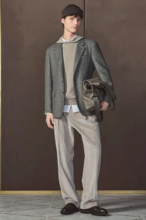 Man in a tweed jacket and corduroy trousers poses indoors holding a bag, showcasing fashion with a warm, neutral color palette.