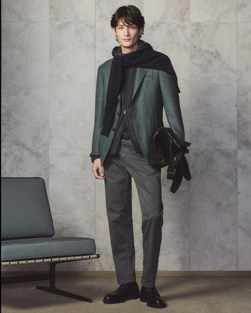 Full-length portrait of an adult male in a blazer, scarf, and trousers, standing indoors against a textured gray marble wall with a chair.
