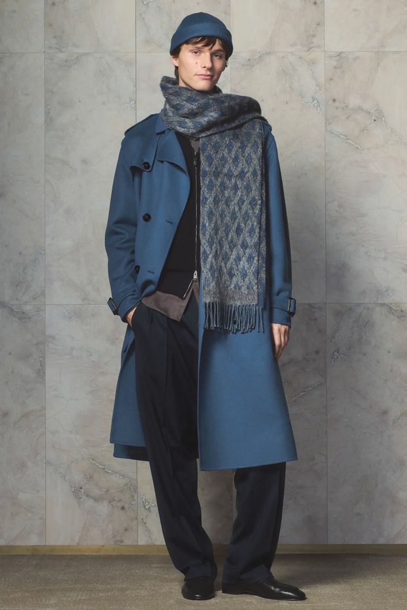 Male model wearing a blue trench coat, scarf, and beanie, posing against a textured wall. Fashion shot with full-body view.