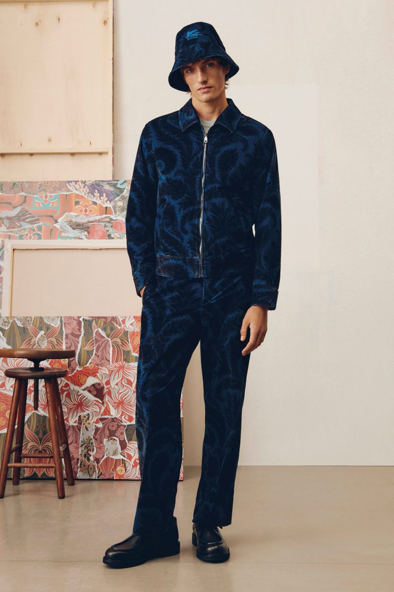A man poses in a blue patterned jacket and pants with a bucket hat against a neutral backdrop with colorful artwork and a stool.