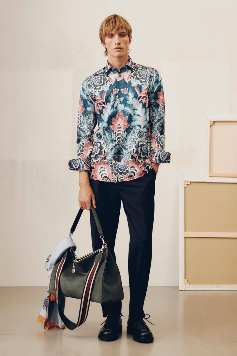 A blond male model in a patterned button-down shirt poses indoors with a shoulder bag, showcasing a fashion-forward, lifestyle aesthetic.