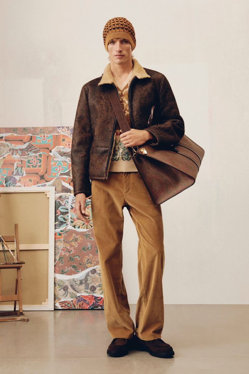 Male model in a brown jacket, tan shirt, and corduroy pants, posing indoors with a brown leather bag and knit hat, fashion editorial.