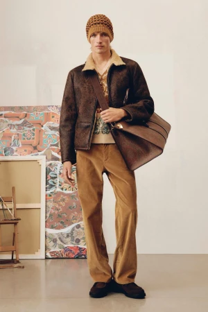 Male model in a brown jacket, tan shirt, and corduroy pants, posing indoors with a brown leather bag and knit hat, fashion editorial.
