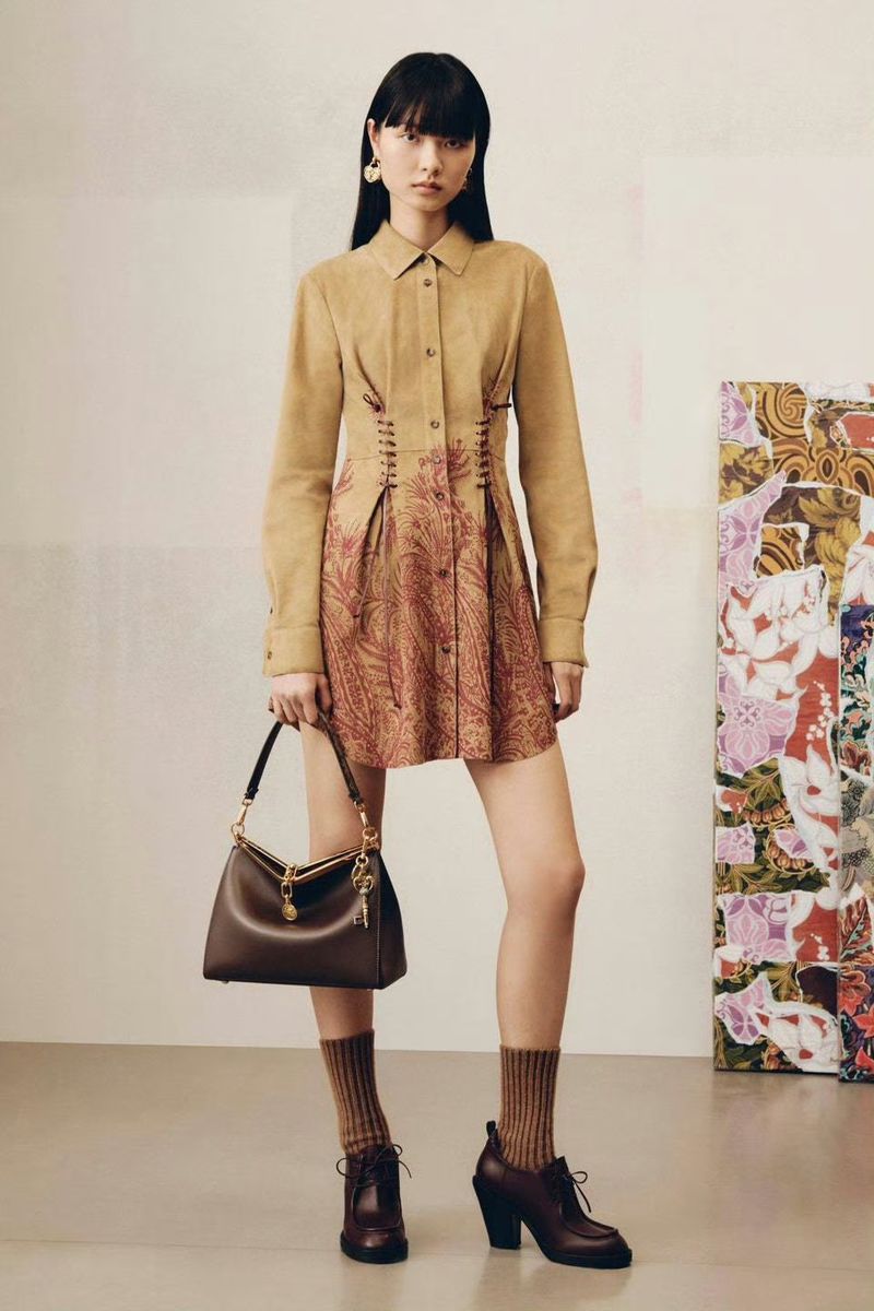 A brunette woman poses indoors wearing a tan long-sleeved dress with brown floral detailing, brown heels, brown socks, and carrying a brown purse.