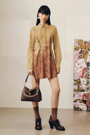 A brunette woman poses indoors wearing a tan long-sleeved dress with brown floral detailing, brown heels, brown socks, and carrying a brown purse.