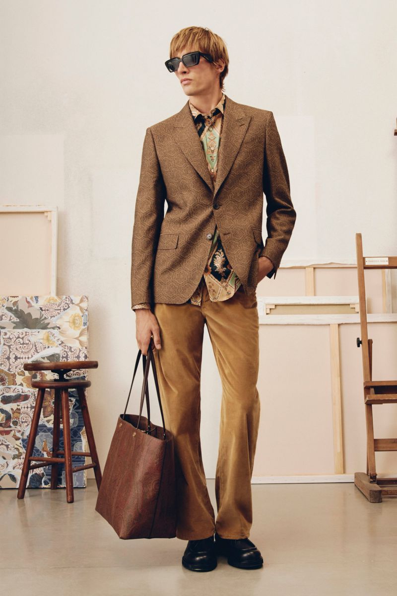 A blond male model in a brown blazer, printed shirt, and flared trousers stands in an art studio, holding a leather tote bag.