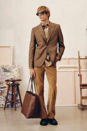 A blond male model in a brown blazer, printed shirt, and flared trousers stands in an art studio, holding a leather tote bag.