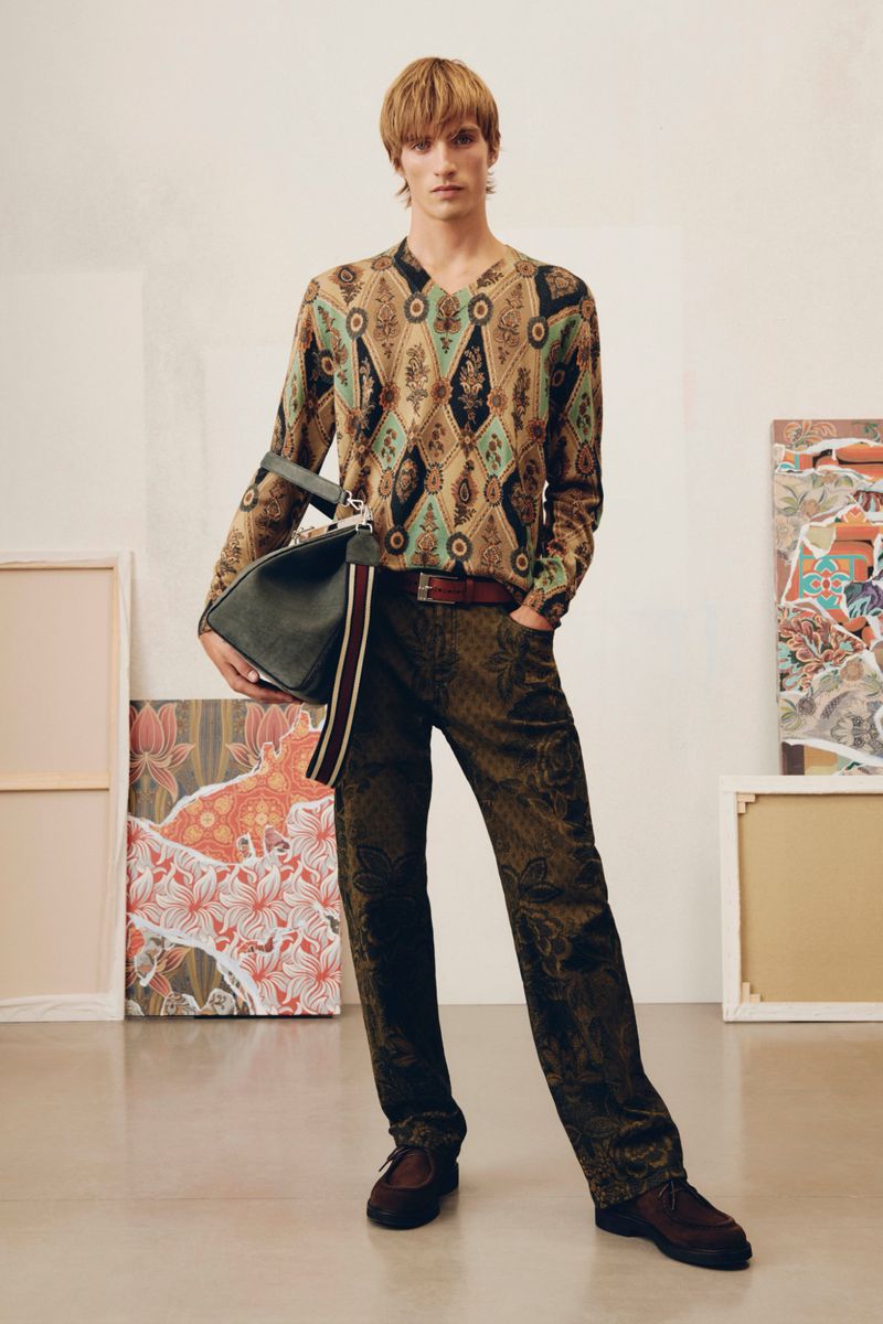 A full-length shot of a male model in a patterned sweater and pants, holding a large leather bag, posing in a photo studio.