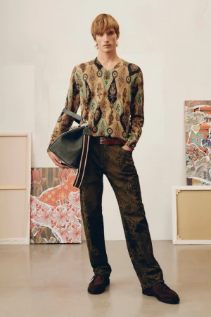 Full shot of a young adult male wearing a patterned shirt and pants, holding a bag, and standing in front of framed artwork.