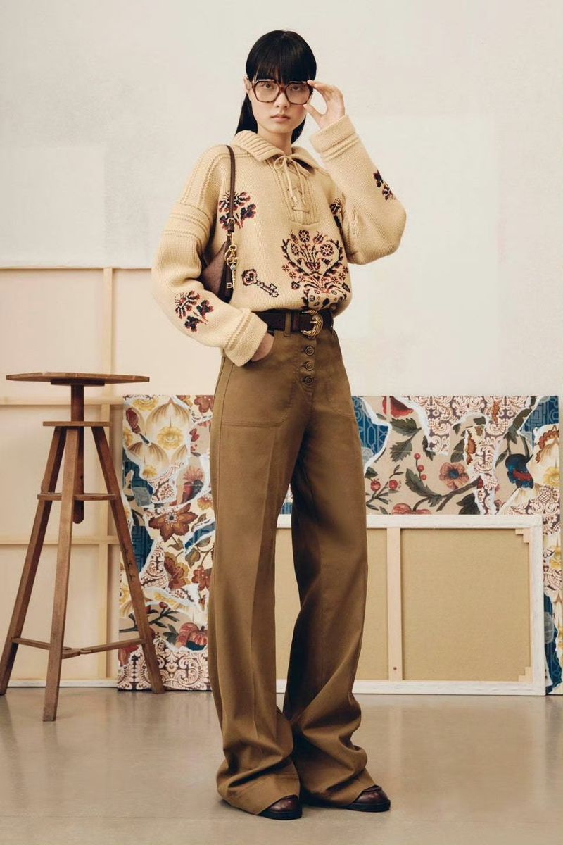 Adult woman models a beige knit sweater with brown floral designs, tan wide-leg pants, and brown shoes indoors with an artful backdrop.