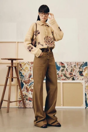 Adult woman models a beige knit sweater with brown floral designs, tan wide-leg pants, and brown shoes indoors with an artful backdrop.