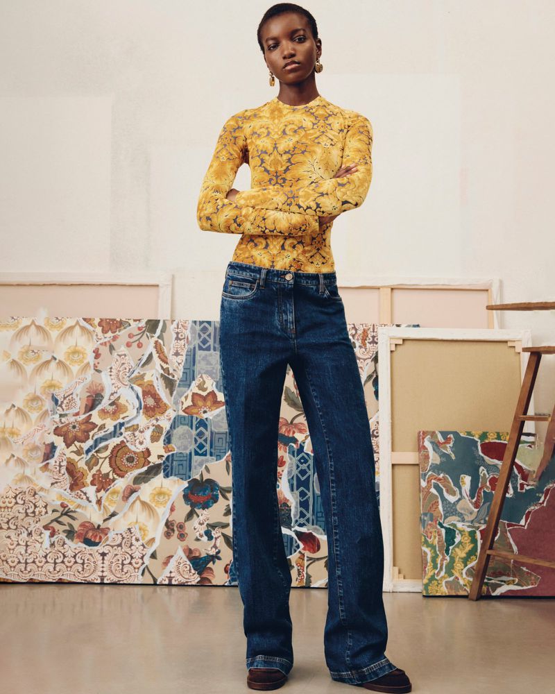 Woman in patterned top and dark denim wide-leg jeans posing in a studio with painted canvas backdrops, fashion editorial shoot.