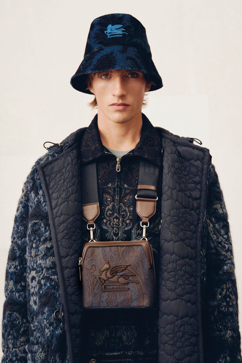 Male model wearing patterned bucket hat, jacket, and shoulder bag, in a studio shot, showcasing contemporary fashion and style.