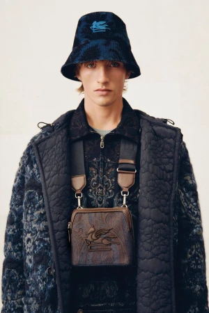 Male model wearing patterned bucket hat, jacket, and shoulder bag, in a studio shot, showcasing contemporary fashion and style.