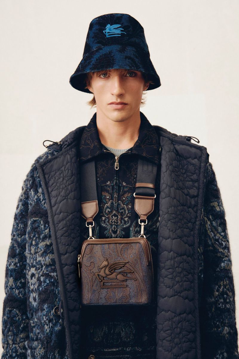 Man in patterned bucket hat and jacket, wearing a crossbody bag with a strap, posing against an off-white backdrop in studio lighting.