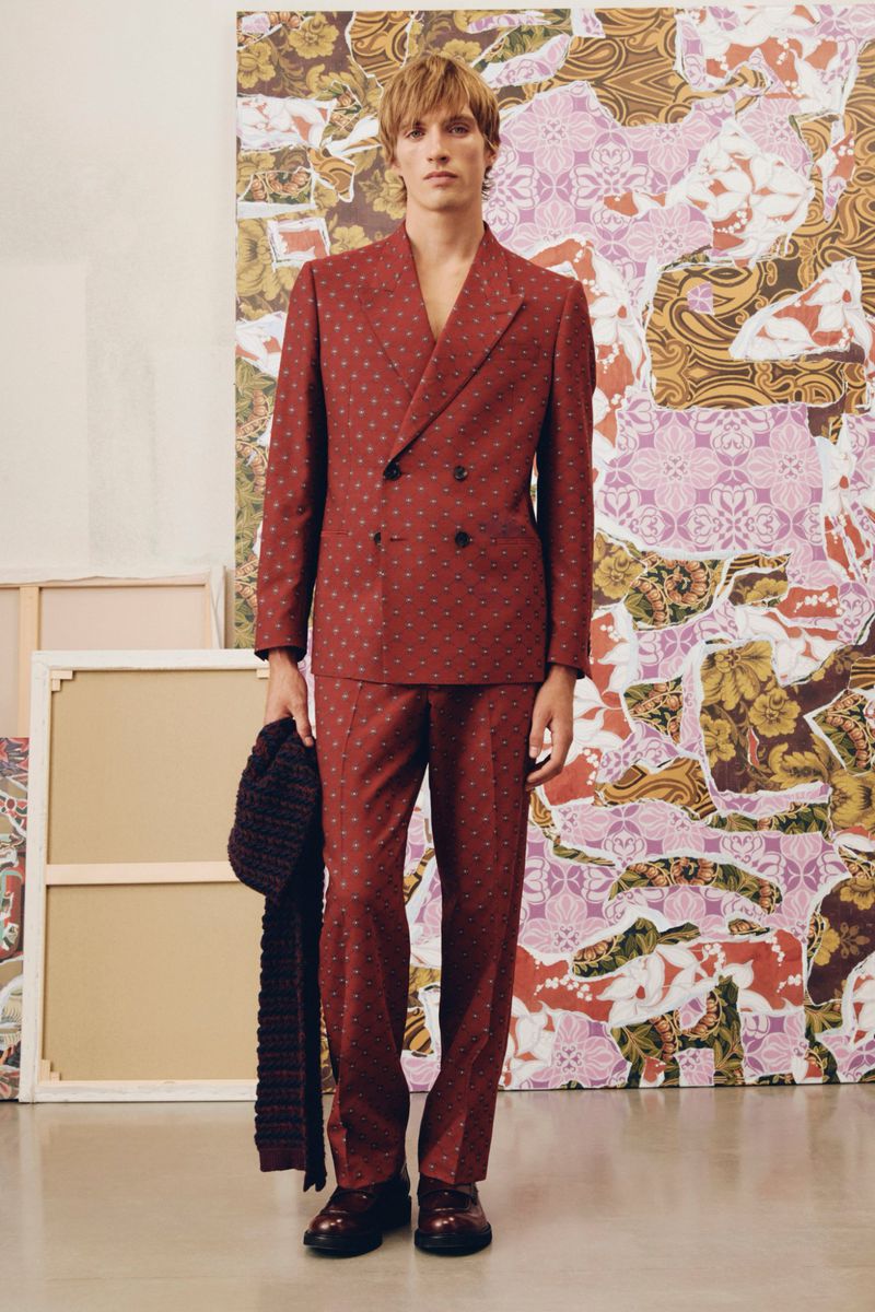 Man in a patterned suit, holding a scarf, stands in front of a painted backdrop and canvases, creating a fashion-editorial look.
