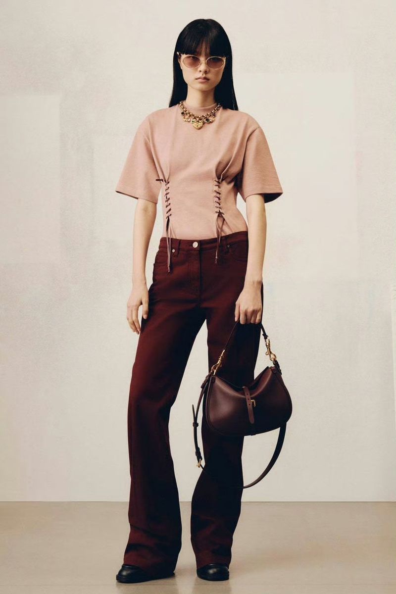 Adult woman in a t-shirt and trousers, holding a handbag in a neutral indoor setting. Fashion editorial image.