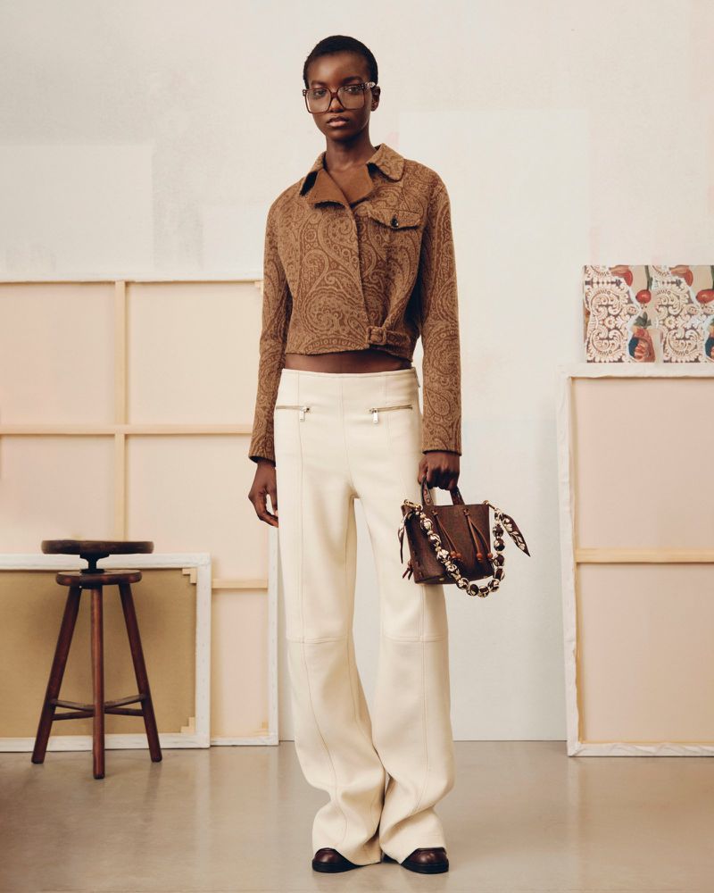 A slender African-American woman models a cropped brown paisley jacket and white pants, complemented by a brown handbag in a studio setting.