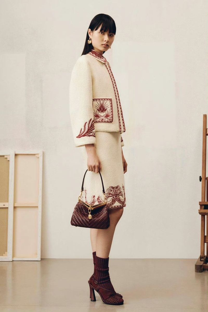 Woman poses in an off-white tweed skirt suit with red floral embroidery, accessorized with a handbag and sock boots in a studio setting.