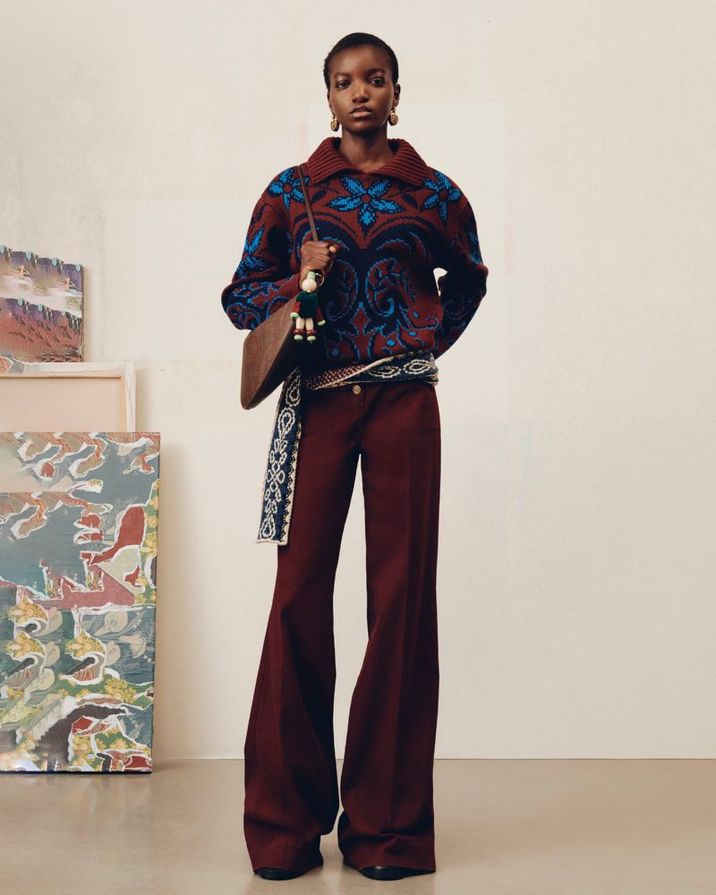 Full-length shot of an African-American woman wearing a patterned sweater, flared pants, and a shoulder bag in a studio setting.