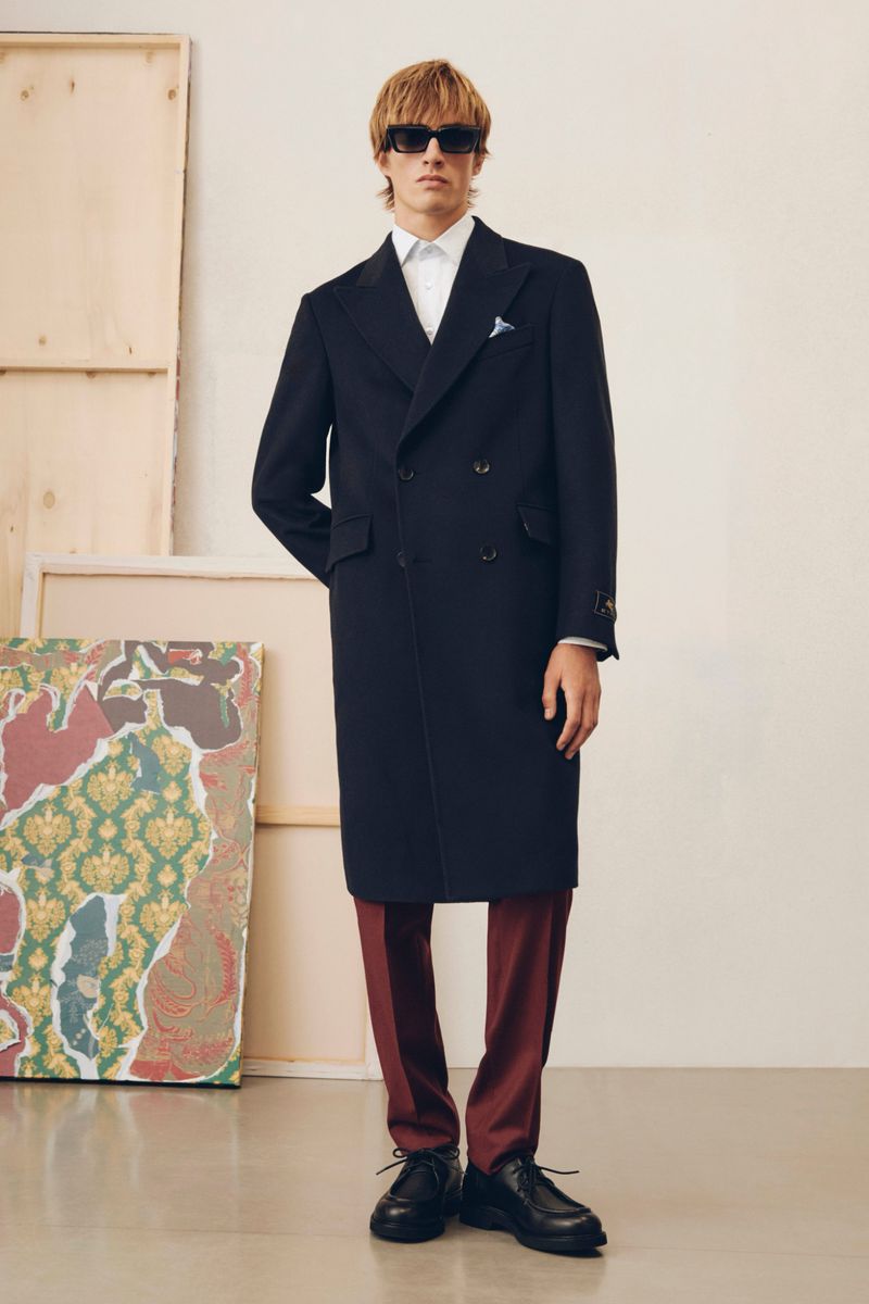 Model in dark sunglasses wearing a dark double-breasted coat and red trousers in a studio with canvas frames