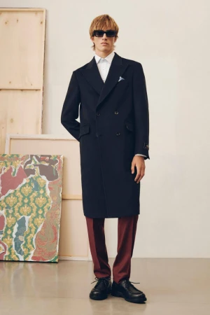 Model wearing a dark double-breasted coat and sunglasses standing in a studio with framed artwork in the background.