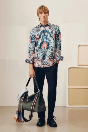 Model wearing a floral patterned button-down shirt holding a green shoulder bag in an indoor studio setting