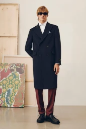 Model wearing a dark double-breasted coat and sunglasses standing in a studio with framed artwork in the background.