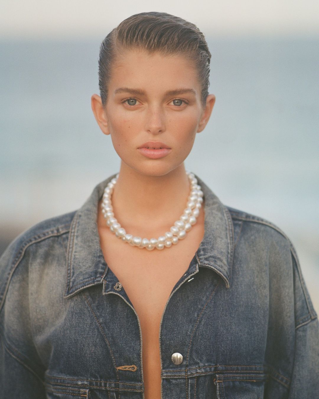 Portrait of a model wearing a denim jacket and a double strand pearl necklace against an outdoor coastal background.