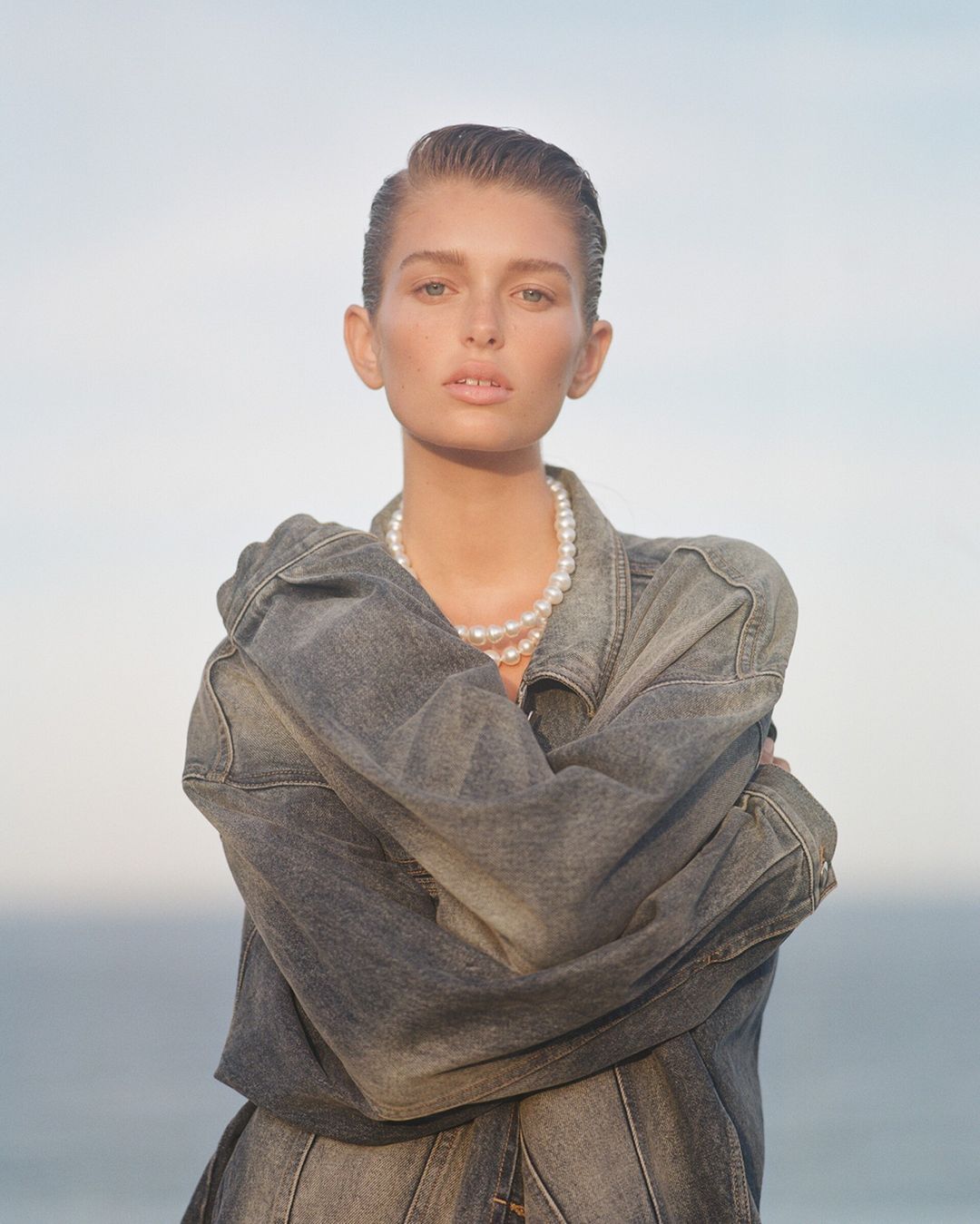 Portrait of a female fashion model with slicked-back hair, wearing a denim jacket and a pearl necklace, posing in front of the ocean.