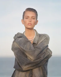Model wearing an oversized denim jacket and pearl necklace against a soft outdoor sky background
