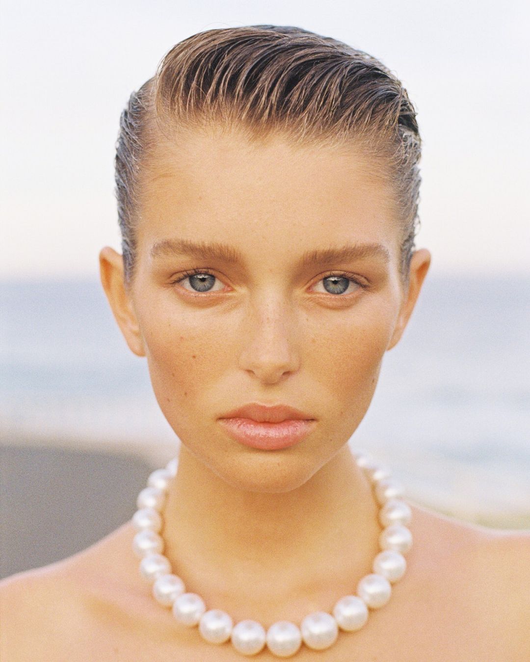 Close-up beauty portrait of a model with slicked-back wet look hair wearing a large pearl necklace against an outdoor coastal background.