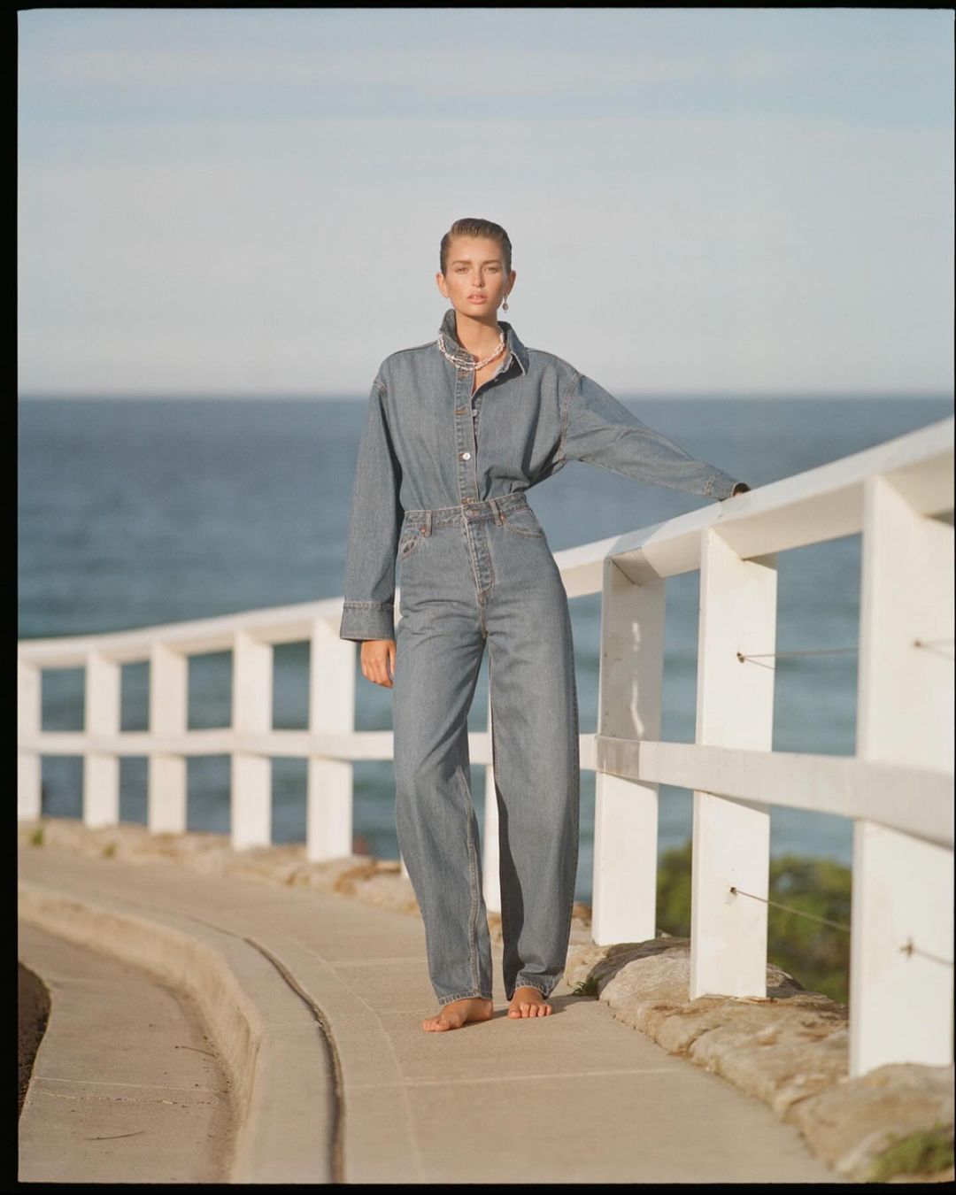Woman poses in denim outfit by the ocean. She leans on a white railing on a concrete path with a blue sky and ocean background.