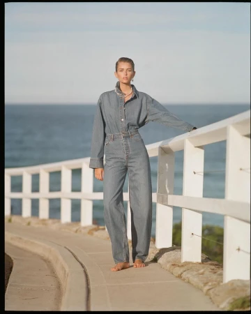 Woman poses in denim outfit by the ocean. She leans on a white railing on a concrete path with a blue sky and ocean background.