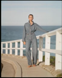 Model standing barefoot on a coastal path wearing a denim jumpsuit, leaning against a white wooden railing by the ocean.