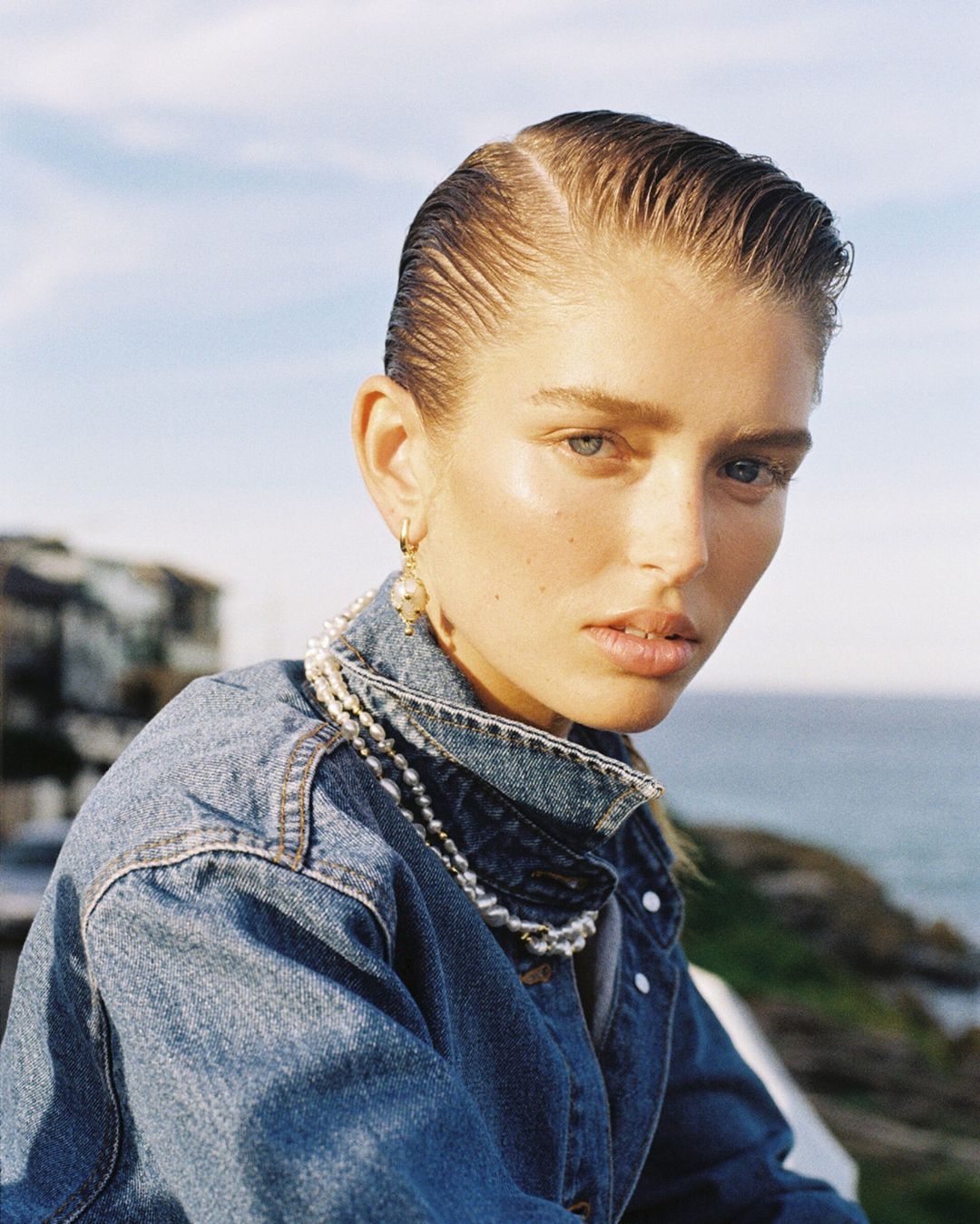 Close-up portrait of a woman with slicked-back hair wearing a denim jacket and pearl necklace against an outdoor coastal background.