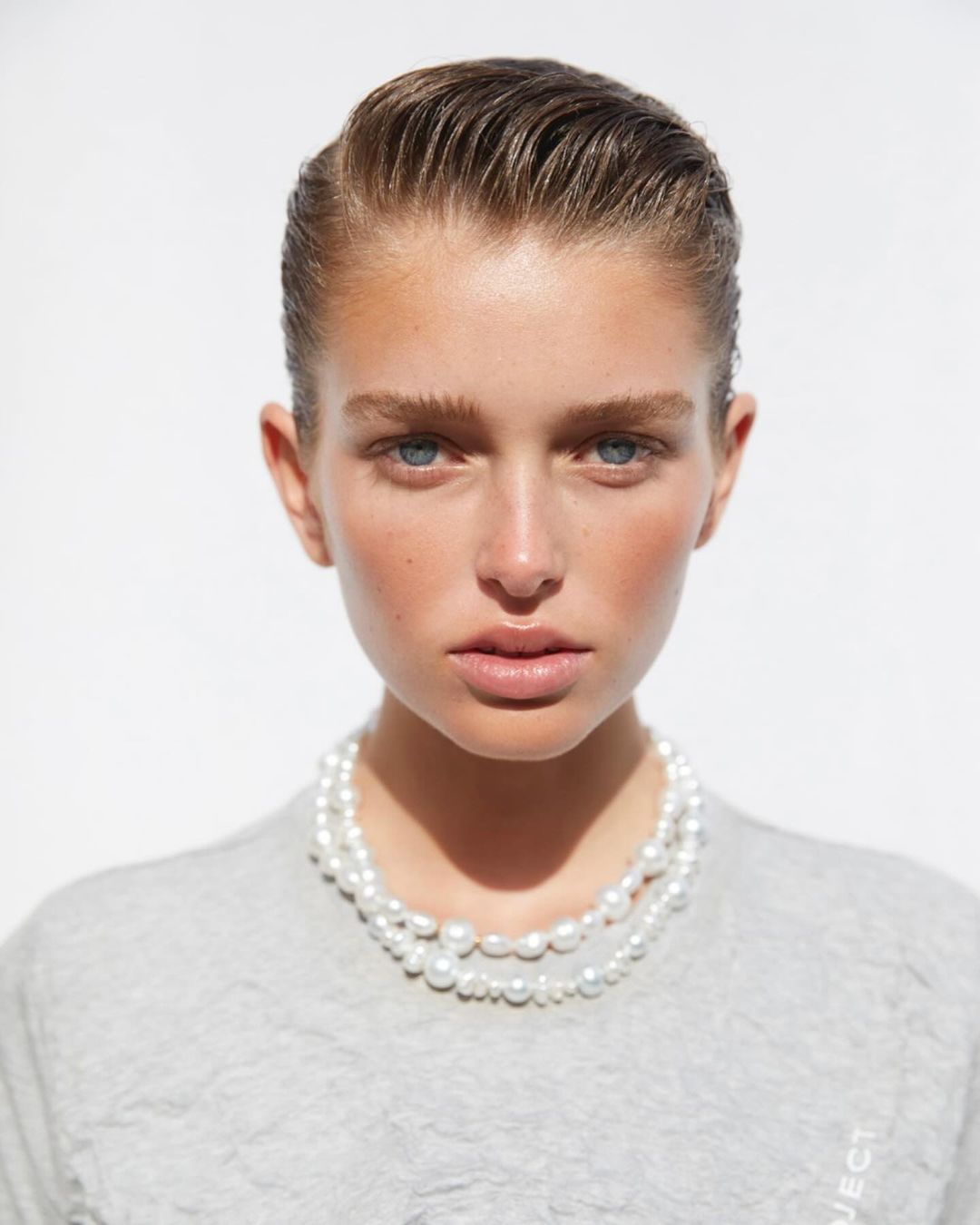 Close-up portrait of a woman with slicked-back hair wearing a double-strand pearl necklace against a bright white background.