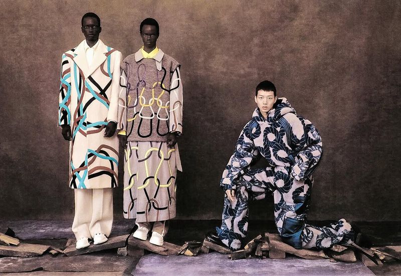 Three models showcase patterned coats and outfits in a studio setting with a brown backdrop, standing on wooden planks.