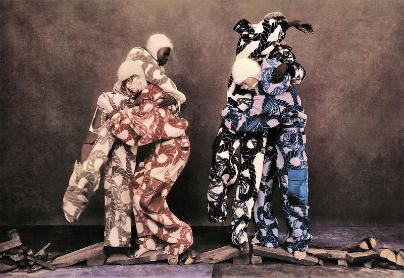 Two pairs of models in avant-garde rope-patterned fashion embrace on a platform stage in a studio with muted colors and dramatic lighting.