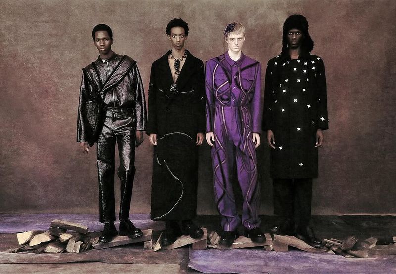 Four adult male fashion models present black and purple apparel in a studio with a textured background and raw wood on the floor.
