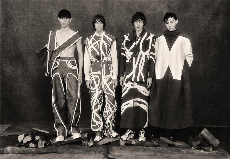 Four Asian models pose in avant-garde fashion against a dark background, standing on wooden planks with piles of bricks and stone.