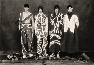 Four male models standing in line wearing patterned dark garments against a textured grey background.
