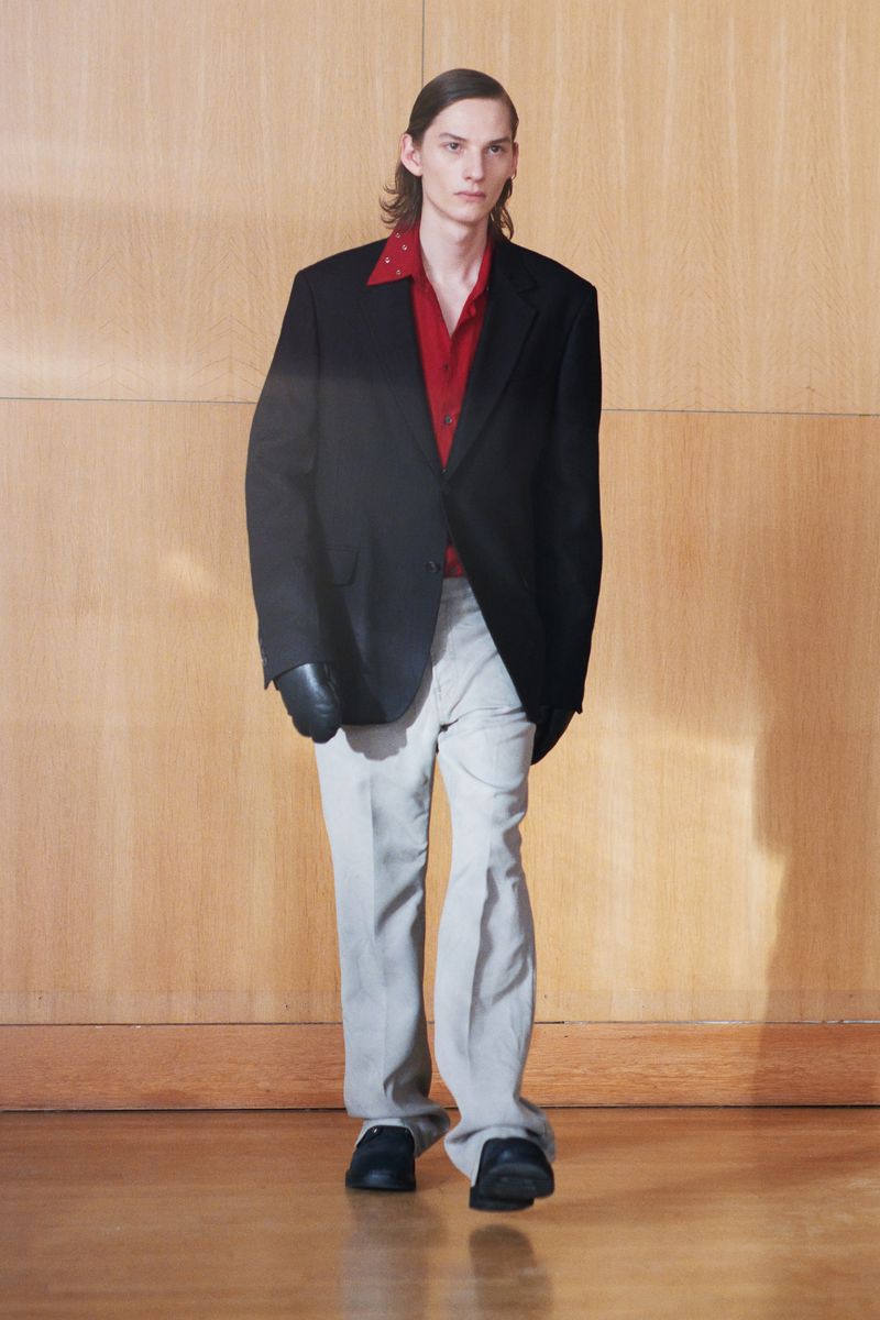 Full-length studio shot of a slender adult male walking forward wearing a black jacket, red shirt, and grey pants in a studio.