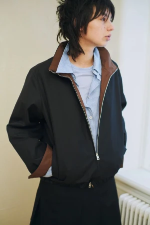 Model wearing a dark jacket with brown trim over a light blue shirt against a plain wall background.