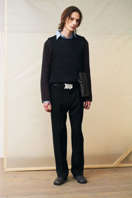 Studio shot of a young adult male model in a black sweater vest, black pants, and silver belt buckle, posing against a neutral backdrop.