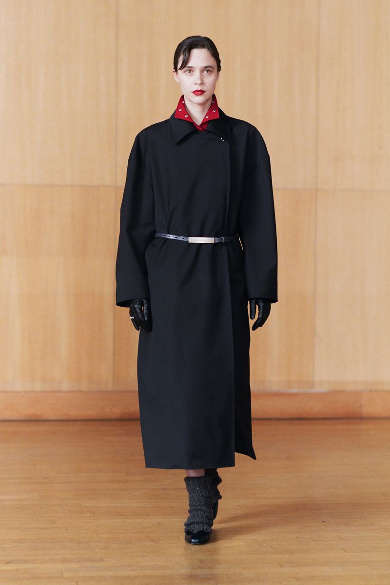 A woman walks toward the camera wearing a black overcoat with a belt, gloves, textured socks, and heels in a minimalist setting.