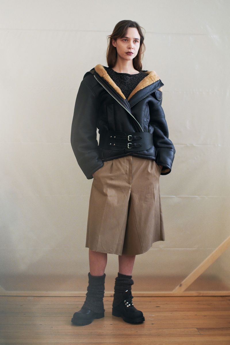 Fashion shot of a woman in a black aviator jacket with fur collar, khaki culottes, and black boots standing indoors in a minimalist setting.