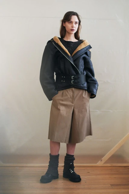 Fashion shot of a woman in a black aviator jacket with fur collar, khaki culottes, and black boots standing indoors in a minimalist setting.
