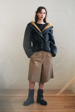 Fashion shot of a woman in a black aviator jacket with fur collar, khaki culottes, and black boots standing indoors in a minimalist setting.