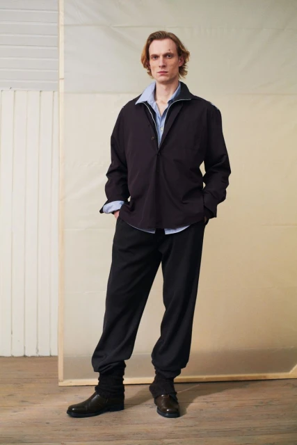 Full shot of an adult male with long hair modelling a black jacket and trousers in a studio with natural light, fashion editorial.